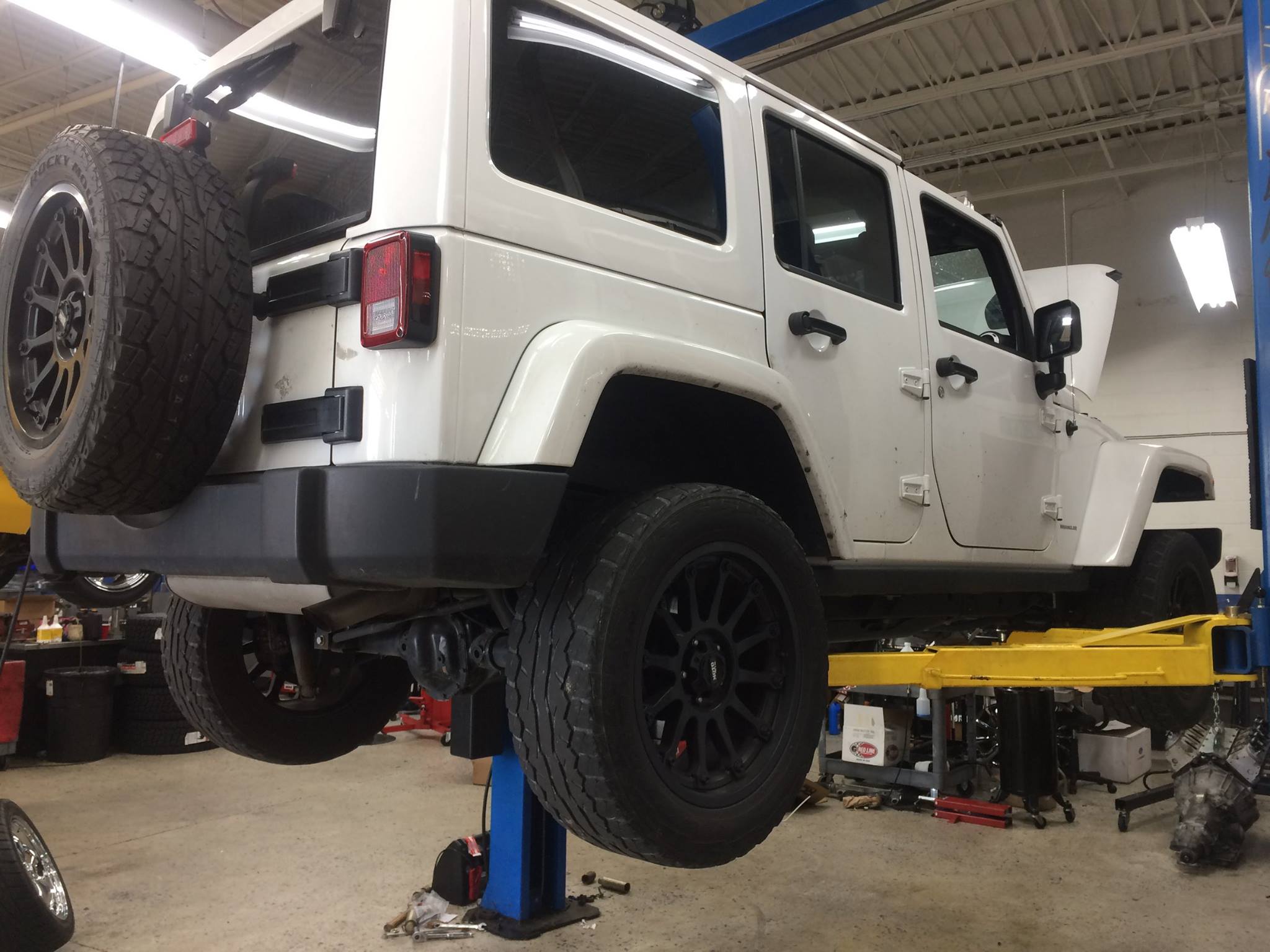 White Jeep Wrangler on a car lift in an auto repair shop, spare tire mounted at the rear and tools on the floor nearby