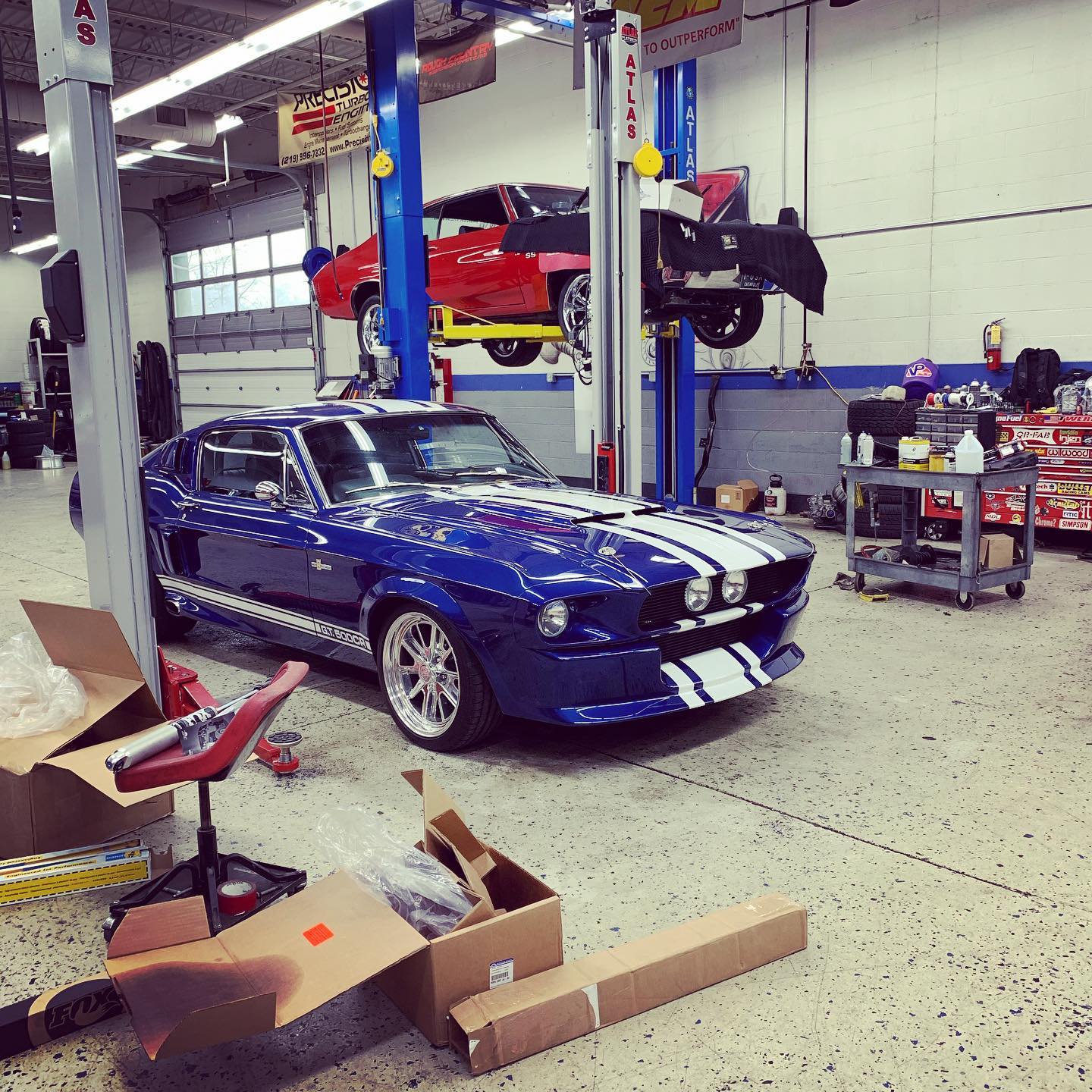 Blue vintage Ford Mustang with white racing stripes parked in a well-equipped auto repair shop near a car lift.