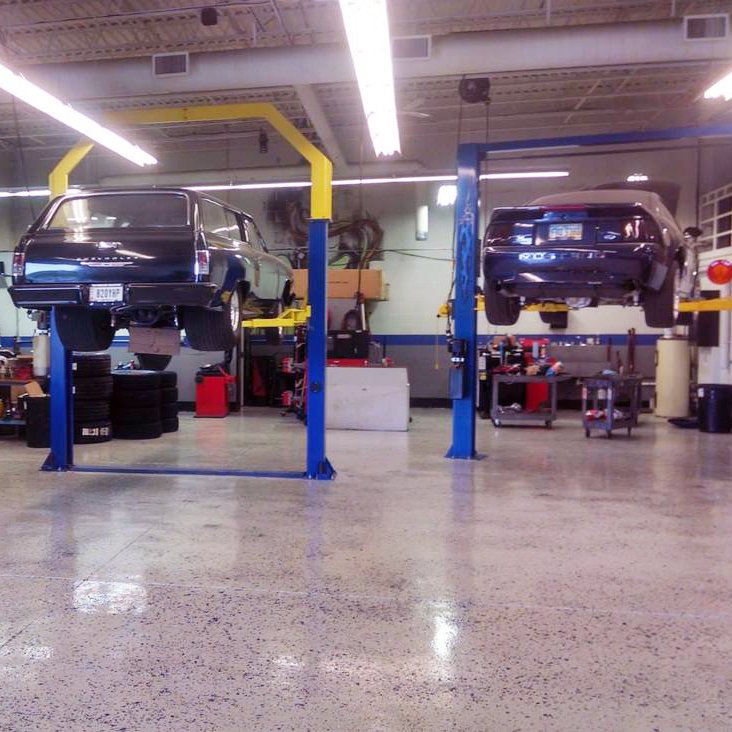 Two vintage cars are raised on hydraulic lifts inside an auto repair shop, with tools and tires in the background.