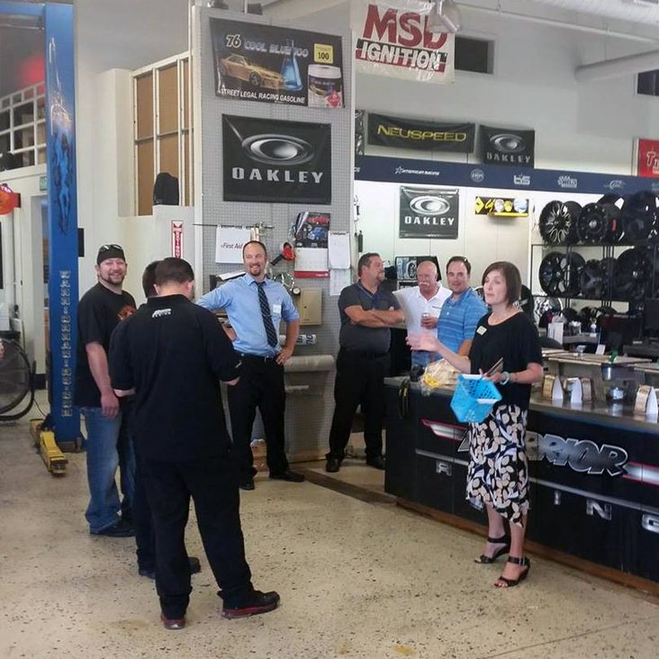 Group of people in a car parts shop gathered and chatting near displays with Oakley banners; a woman holds a blue basket at the foreground.