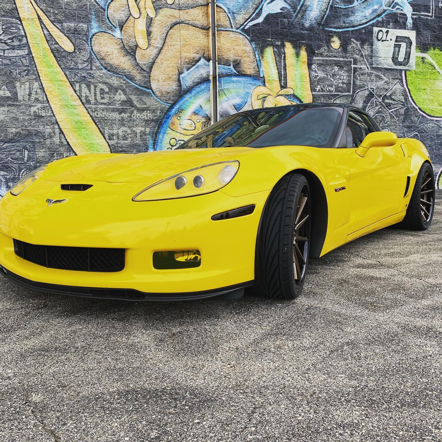 Yellow Chevrolet Corvette sports car parked in front of a colorful graffiti wall, view of front-left side and open top shade area visibleed, capturing sleek lines and bright color.